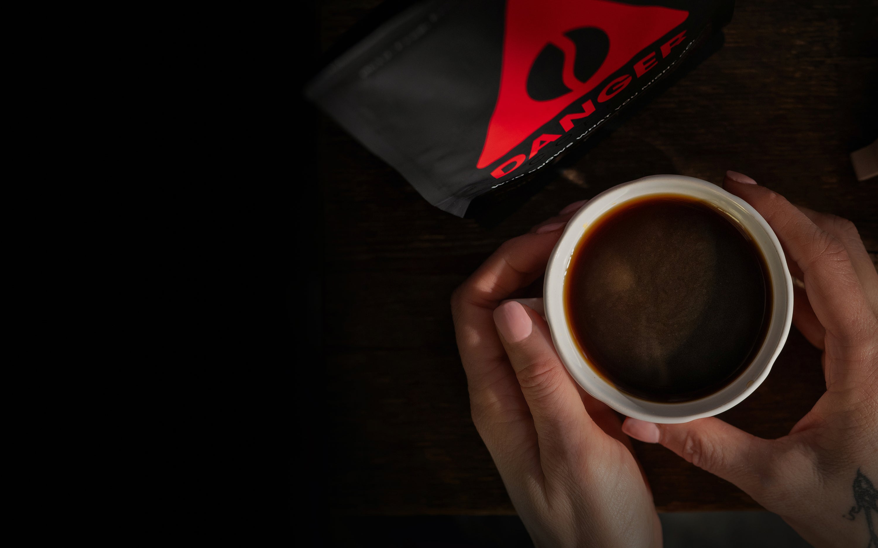 Overhead shot of hands holding black coffee beside a Danger Coffee bag with red triangle logo.
