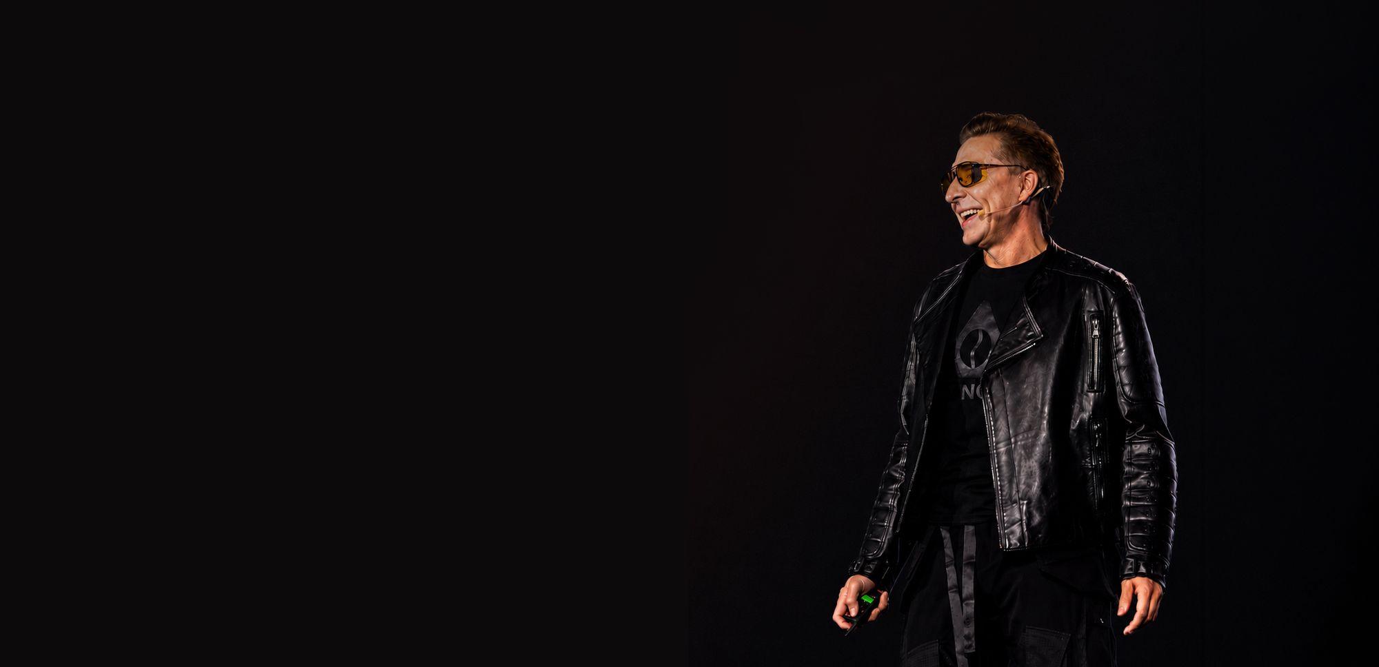 A man wearing a black leather jacket, black t-shirt, and sunglasses stands against a solid black background, looking to the left with a slight smile.