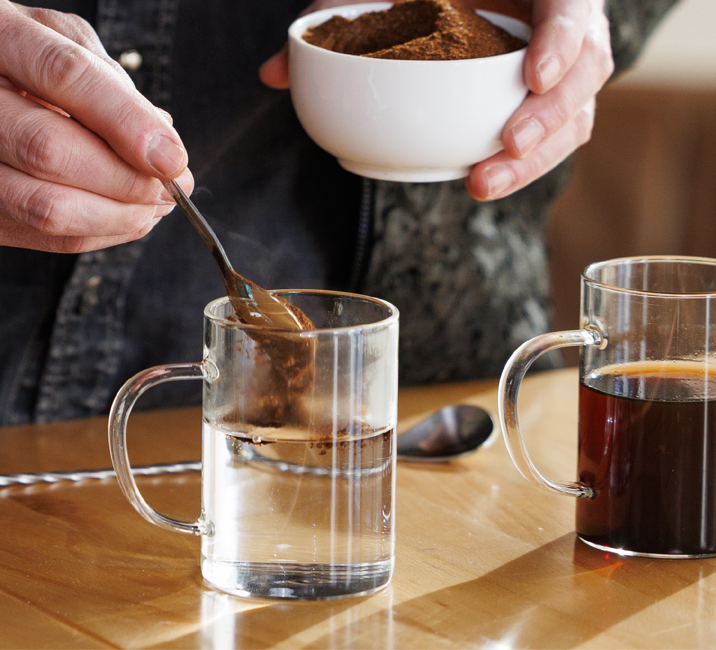 Person stirring a clear mug with a spoon, holding a white bowl of Instant Danger on a wooden table.
