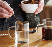 Person stirring a clear mug with a spoon, holding a white bowl of Instant Danger on a wooden table.