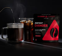 Glass mug with steaming coffee, bag of instant coffee powder, and 'Danger Coffee' packaging on a dark background.
